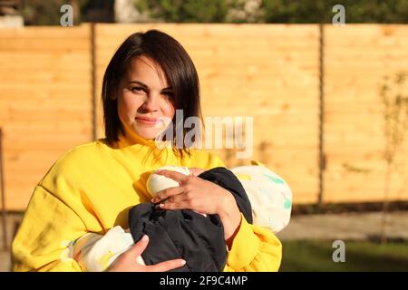 Une belle jeune mère avec des cheveux bruns droits et dedans un sweat-shirt jaune tient un petit enfant dans ses bras dans le parc dans les rayons d'un coucher de soleil Banque D'Images