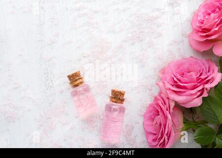 Deux bouteilles d'huile de rose, cristaux de sels minéraux pour le bain et roses roses roses sur la table en bois blanc. Banque D'Images