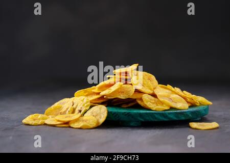 Croustilles de banane séchées ou gaufres de banane, éparpillées sur une base ronde verte avec fond texturé gris, isolées. Banque D'Images