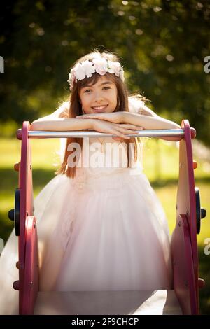 Belle fille caucasienne vêtue pour la communion dans le parc haut d'une diapositive regardant l'appareil photo Banque D'Images