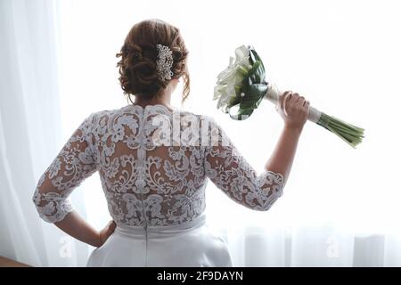 Vue arrière d'une superbe mariée tenant son bouquet d'attente pour son marié en se tenant près de la fenêtre Banque D'Images
