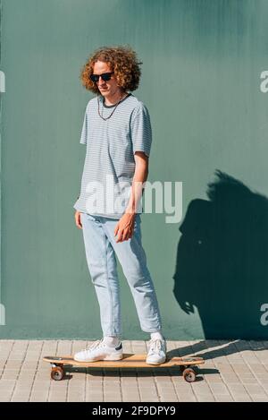 portrait vertical d'un jeune skateboarder à bord de son skate sur un fond vert. Il a de longs cheveux bouclés et porte des lunettes de soleil Banque D'Images
