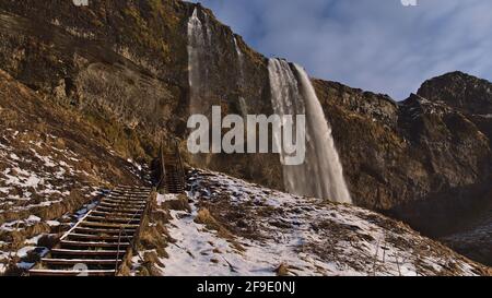 Vue imprenable sur la célèbre chute d'eau Seljalandsfoss sur la côte sud de l'Islande près du périphérique le jour d'hiver ensoleillé avec des escaliers en bois. Banque D'Images