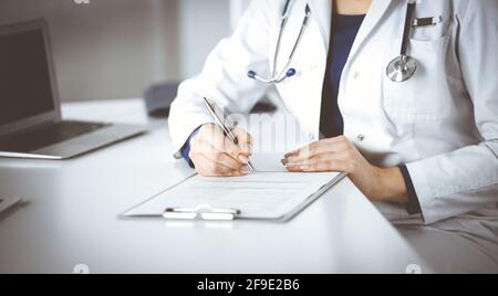 Une femme-médecin inconnue prescrit des médicaments à son patient, à l'aide d'un presse-papiers, tout en étant assise sur le bureau de son cabinet. Femme médecin Banque D'Images