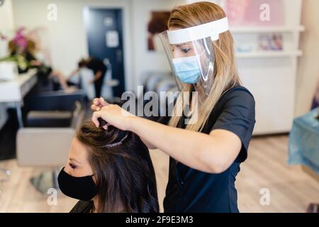 Coiffeur, protégé par un masque, peignant les cheveux de son client avec un fer à repasser dans un salon. Banque D'Images