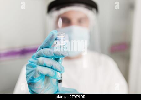 Médic flou non reconnaissable dans le masque de protection du visage et le latex gants avec flacon de vaccin contre le coronavirus et seringue montrant à appareil photo en mode stan Banque D'Images