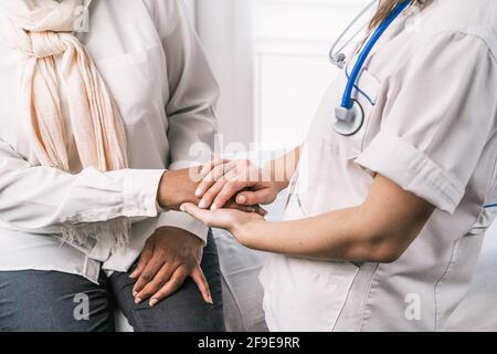 Femme non reconnaissable cultivée médecin en uniforme médical et stéthoscope portant un masque parlant et tenant les mains d'une femme africaine américaine mature Banque D'Images