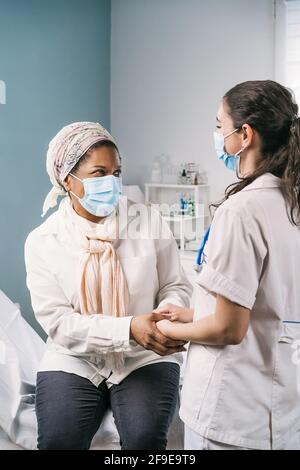 Jeune femme médecin en uniforme médical et stéthoscope portant un masque facial parlant et tenant les mains de la femme africaine américaine mature pendant l'ap Banque D'Images