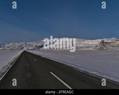 Perspective décroissante étonnante de la rocade pavée déserte (route 1) avec des marquages sur la route dans le sud de l'Islande en hiver avec les montagnes. Banque D'Images