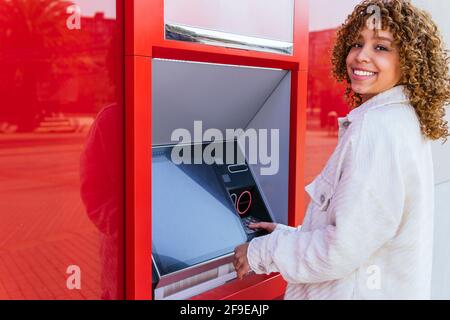 Vue latérale d'une femme afro-américaine souriante à l'aide d'un guichet automatique et retirer de l'argent en se tenant dans la rue de la ville appareil photo Banque D'Images