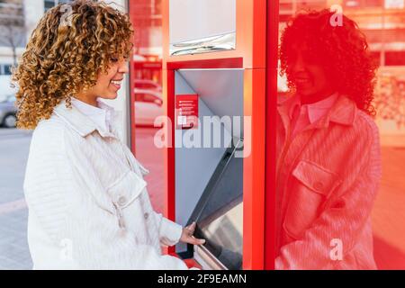 Vue latérale d'une femme afro-américaine souriante à l'aide d'un guichet automatique et retirer de l'argent en se tenant dans la rue de la ville Banque D'Images