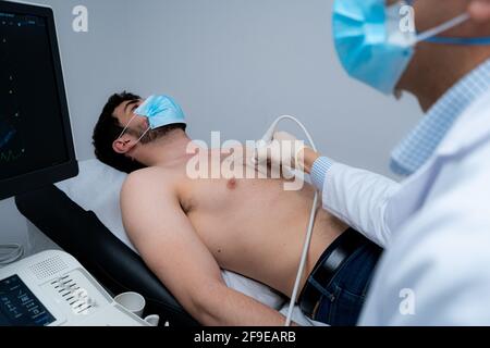 Vue latérale d'un médic masculin non reconnaissable court à l'aide d'une échographie moderne l'équipement et l'examen du cœur d'un patient masculin dans le masque couché sur table médicale du Banque D'Images