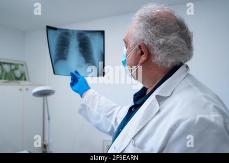 Vue latérale d'un médecin de sexe masculin âgé avec masque et gants examen de l'image radiographique des poumons du patient pendant le coronavirus à l'hôpital Banque D'Images