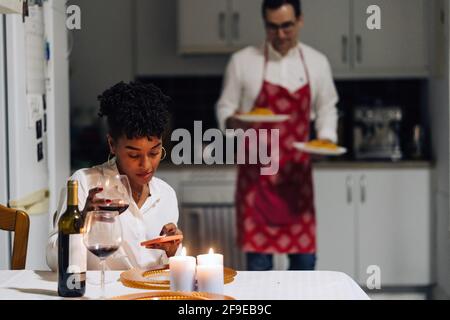 Femme noire assise à la table et parcourant un smartphone pendant que l'homme sert de la nourriture pour un dîner romantique à la maison Banque D'Images