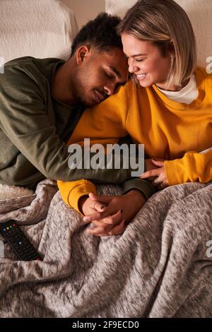 Grand angle d'amour multiethnique couple se relaxant sur le canapé sous couvrir tout en se coulant et en tenant les mains Banque D'Images