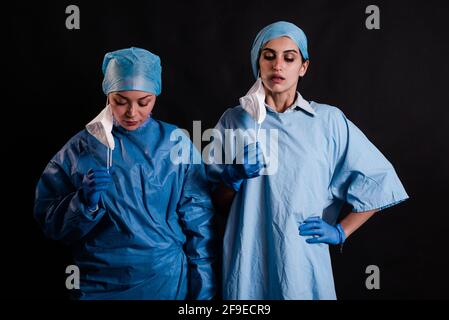Jeunes femmes collègues en uniformes médicaux qui prennent des masques faciaux en se tenant sur fond noir en clinique Banque D'Images