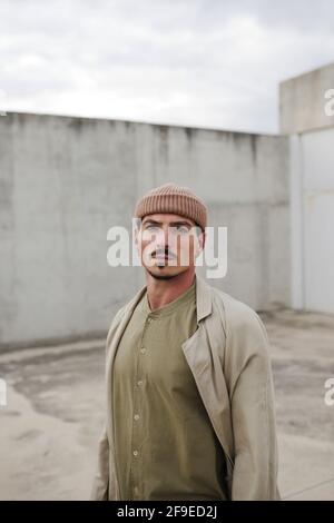 Homme sérieux portant un manteau et un chapeau tendance debout en ville et regarder l'appareil photo Banque D'Images