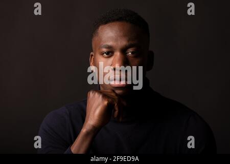 Jeune homme afro-américain confiant, vêque de noir à la main au menton regardant l'appareil photo sur fond noir en lumière studio Banque D'Images