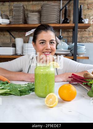 Femme joyeuse regardant l'appareil photo à la table avec un pot de délicieux smoothie végétalien et agrumes frais dans la cuisine Banque D'Images