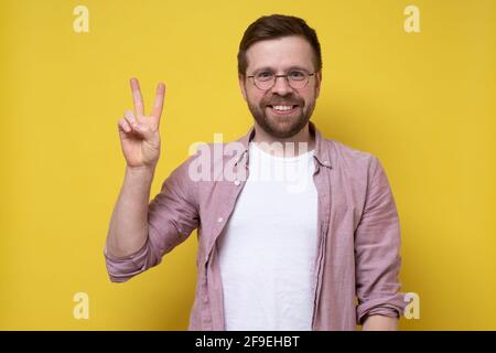 Joyeux homme en lunettes et vêtements décontractés montre le symbole de la victoire et regarde la caméra sourire. Chiffre avec deux doigts. Banque D'Images