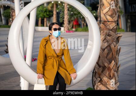 Femme solitaire asiatique sur une chaise pivotante à l'extérieur portant le visage masque et distance sociale en public Banque D'Images