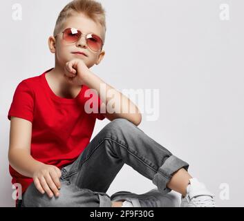 Gai élégant garçon en t-shirt rouge, jeans, baskets et lunettes de soleil assis sur le sol tenant le menton avec la main regardant l'appareil photo Banque D'Images