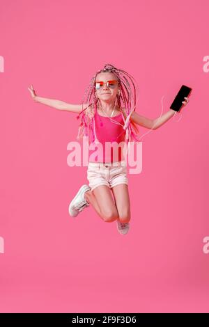 Belle petite fille avec des dreadlocks sautant avec le téléphone mobile dedans la main et écoutez de la musique sur un casque sur fond rose Banque D'Images