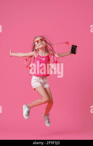 Belle petite fille avec des dreadlocks sautant avec le téléphone mobile dedans la main et écoutez de la musique sur un casque sur fond rose Banque D'Images