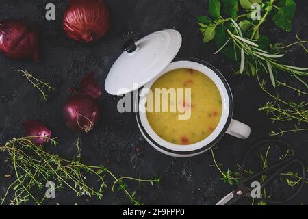 Vue de dessus de la soupe de pommes de terre dans un bol blanc avec poignée et couvercle sur le fond arrière, décoré avec des oignons rouges et des herbes Banque D'Images