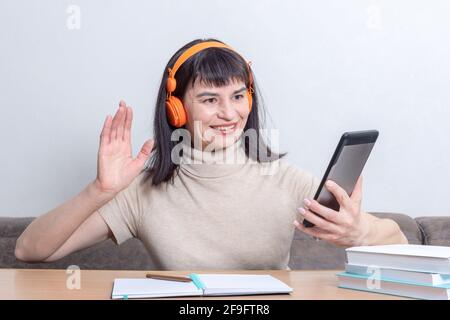 Charmante femme brune dans un casque orange avec un appel vidéo et un geste de salutation tout en étant assis à la table à la maison. Utilisation d'un appareil électronique portable Banque D'Images