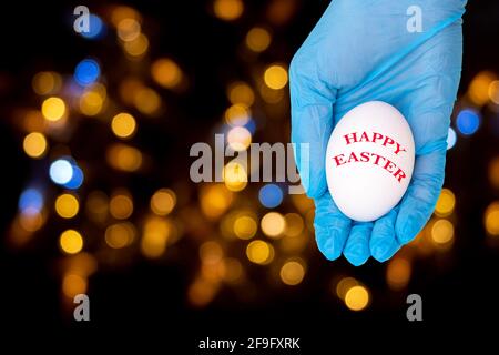 Une main dans un gant médical de protection tient ou donne un oeuf blanc avec l'inscription JOYEUSES PÂQUES sur un fond noir avec bokeh, espace de copie. Heureux Banque D'Images