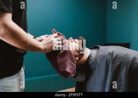 Le coiffeur se nettoie à l'aide d'une serviette sur la tête d'un client mâle barbu qui est allongé dans une chaise de salon de coiffure. Banque D'Images