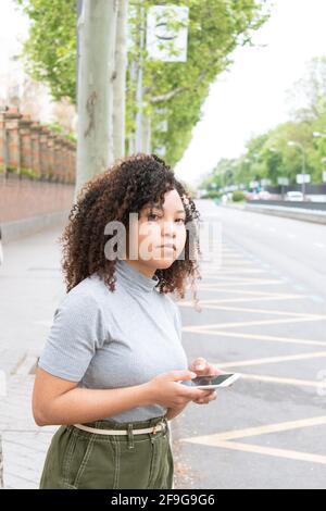 jeune fille aux cheveux bouclés utilisant un téléphone cellulaire en attendant un taxi ou uber dans la rue. Photo verticale. Banque D'Images
