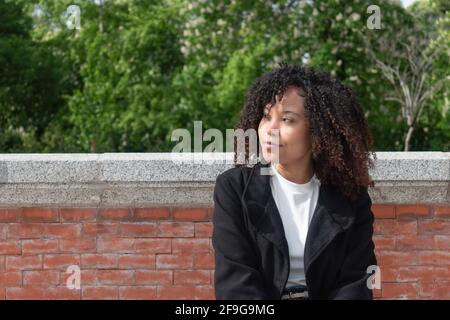 Jeune femme avec des cheveux bouclés regardant hors de la vue de la gauche avec un fond d'arbres et de briques. Banque D'Images