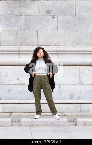 Jeune femme aux cheveux bouclés regardant directement dans l'appareil photo. Veste noire et pantalon vert. Photo verticale. Banque D'Images