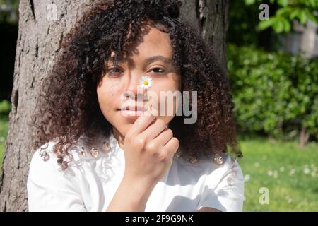 Jolie jeune femme aux cheveux bouclés regardant l'appareil photo tout en tenant une petite fleur devant son visage Banque D'Images