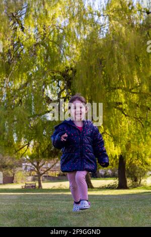 Photo pleine longueur d'une petite fille à cheveux bruns mignonne avec veste bleue et pantalon rose sur l'herbe dans un parc par un beau jour de printemps Banque D'Images