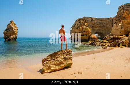 Jeune homme debout sur un rocher, plage de Praia da Marinha, côte rocheuse, formation rocheuse dans la mer, Algarve, Lagos, Portuga Banque D'Images