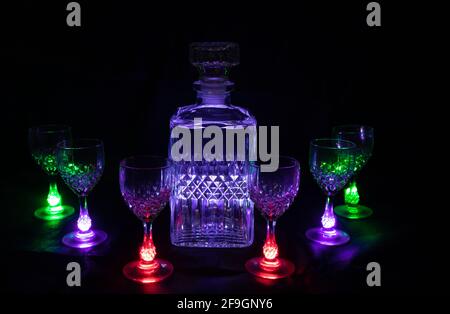 Une bouteille de liqueur en cristal vide avec six verres en cristal éclairés avec des lumières de différentes couleurs sur fond noir Banque D'Images