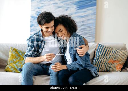 Concept de parenthood, attente de bébé, test de grossesse positif. Joyeux couple multiracial s'asseoir et embrasser sur le canapé à la maison et regarder le test de grossesse positif, aller avoir un enfant, heureux au sujet de bonnes nouvelles Banque D'Images