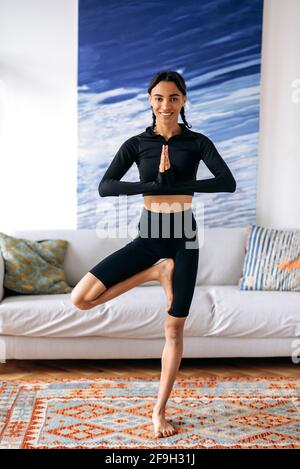 Belle jeune femme afro-américaine en vêtements de sport, mène un style de vie sportif, s'étirant à la maison, debout dans la posture de l'arbre, regarde l'appareil photo, souriant amical. Style de vie sain et sportif Banque D'Images