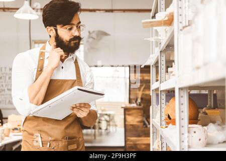 Jeune homme d'affaires séduisant un potter avec une barbe et une moustache travaille dans son atelier. Conserve les enregistrements et transcrites dans un ordinateur portable en inspectant Banque D'Images