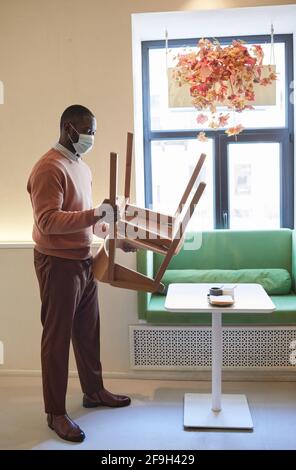 Portrait complet d'un homme afro-américain portant un masque dans un café et arranger les meubles tout en préparant l'ouverture le matin Banque D'Images