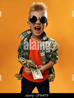 Un petit garçon en lunettes de soleil rondes, une chemise à carreaux et un Jean avec un lot de dollars en mains et des cris Banque D'Images