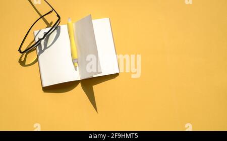 Ouvrez le bloc-notes, le stylo et les lunettes de lecture sur le dessus du tableau de fond jaune. Vue de dessus, espace de copie. Banque D'Images