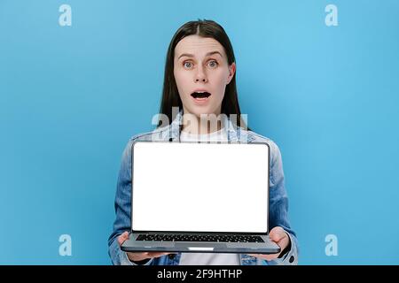 Une jeune femme excitée se tenant sur un ordinateur portable, choquée de regarder l'appareil photo, vêtue d'une veste en denim, posant isolée sur un mur bleu d'arrière-plan de studio Banque D'Images