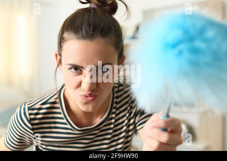 Vue de face d'une drôle de femme grimaquant menaçant à un appareil photo avec un nettoyant duster à la maison Banque D'Images