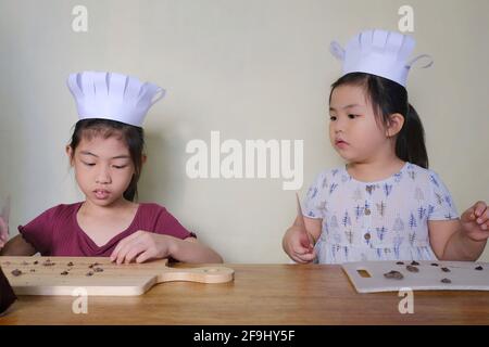 Deux jeunes sœurs asiatiques mignonnes, avec de jolis chapeaux de chef, apprennent à fabriquer des biscuits aux pépites de chocolat, en coupant du chocolat en petits morceaux avant de mixer Banque D'Images