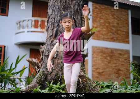 Une jeune fille asiatique mignonne est en train de marcher et de se balancer sur la racine d'un grand arbre dans son arrière-cour, en s'amusant et souriant. Banque D'Images