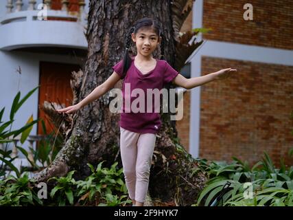 Une jeune fille asiatique mignonne est en train de marcher et de se balancer sur la racine d'un grand arbre dans son arrière-cour, en s'amusant et souriant. Banque D'Images